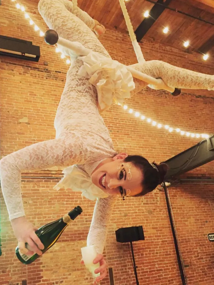 Aerialist hanging upside down and pouring champagne at a reception under string lights