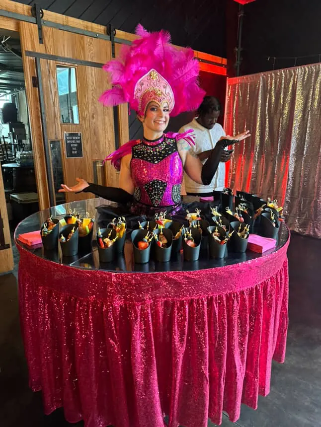 Strolling table performer in a pink feathered headdress serving champagne from a table skirt